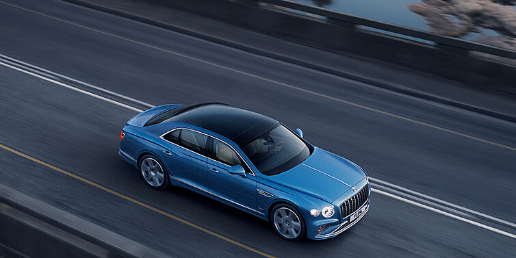 Bentley Lugano New Bentley Flying Spur Azure sedan in Light windsor blue paint by Mulliner, with 22 inch Azure wheels seen from overhead driving dynamically on a country road