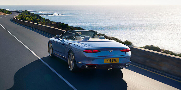 Bentley Lugano New Bentley Continental GTC Azure convertible rear view in Light Windsor blue paint with 22 inch ten spoke wheels driving dynamically on an ocean road in the sunshine